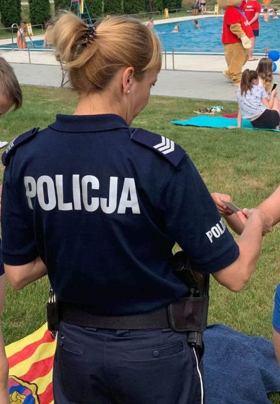 Fotografia kolorowa. Na zdjęciu widoczna policjantka stojąca tyłem wręczająca ulotki dzieciom.
