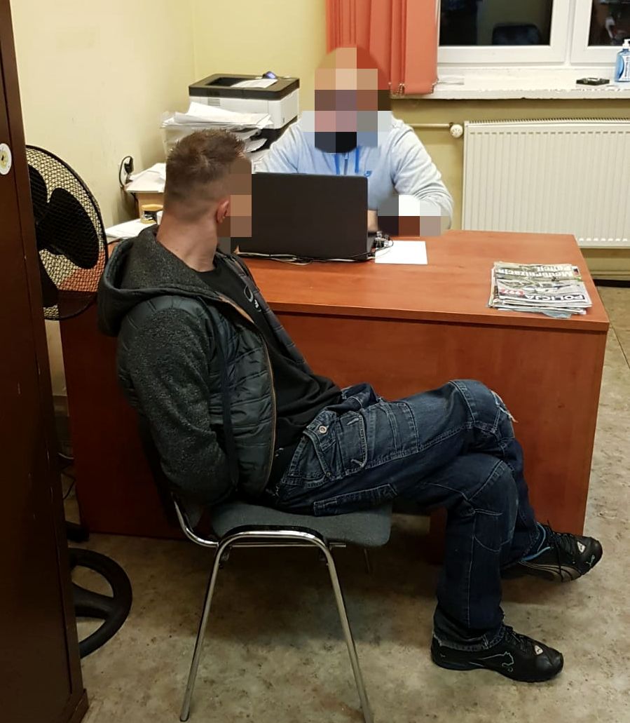 Na zdjęciu widoczny policjant siedzący przy biurku oraz siedzący na krześle zatrzymany
