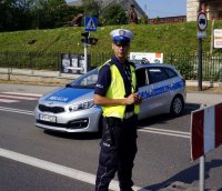 Fotografia kolorowa. Na zdjęciu policjant w odblaskowej kamizelce stojący przy radiowozie, zabezpieczający skrzyżowanie.