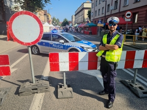 Zdjęcie przedstawia policjanta stojącego przy barierkach ochronnych. W tle zamknięta droga i idący nią pielgrzymi